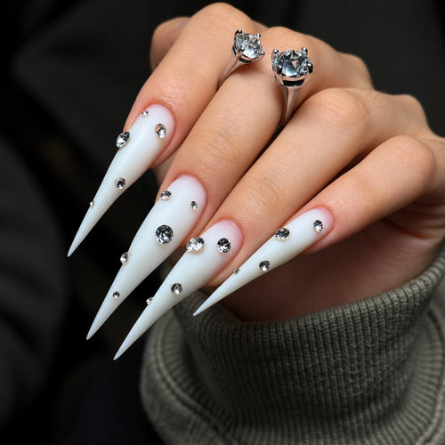 Extra-long matte white claw press-on nails with sparkling rhinestone accents, displayed on a hand against a dark background.